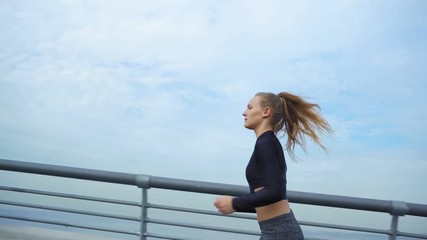 Tracking left of fit female jogger with ponytail running on bridge road over water during marathon training, side view slow motion shot