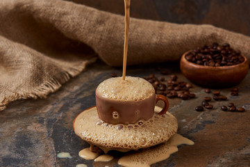 flowing out hot coffee in cup with foam and puddle on saucer on marble surface near sackcloth and beans