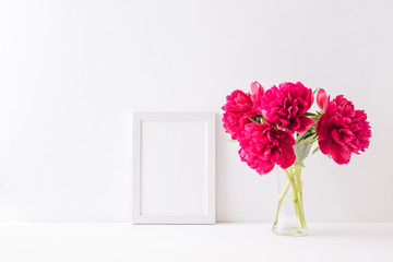 Mockup with a white frame and red peonies in a vase on a white table