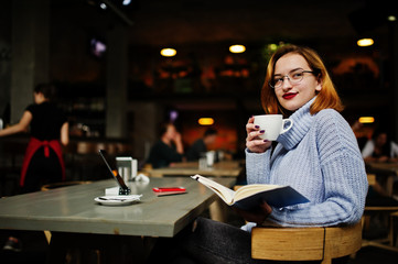 Cheerful young beautiful redhaired woman in glasses using her phone, touchpad and notebook while sitting at her working place on cafe with cup of coffee.