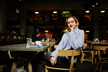 Cheerful young beautiful redhaired woman in glasses sitting at her working place on cafe with cup of coffee.