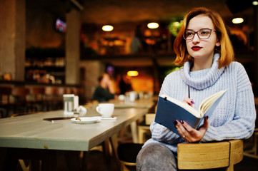 Cheerful young beautiful redhaired woman in glasses sitting at her working place on cafe reading something at her notebook.