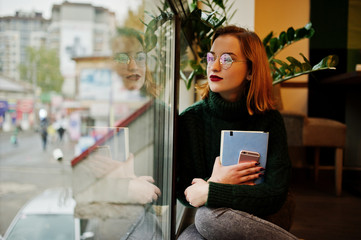 Cheerful young beautiful redhaired woman in glasses, green warm wool sweater, using her notebook, while sitting on cafe against window.