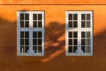 Two old style white wooden windows on a bright orange wall, partly covered by a shadow.