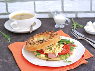 Breakfast, bagel with scramble egg, ham, arugula and vegetables on a white plate on a dark background.