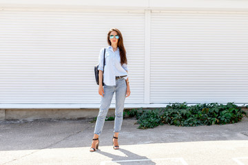 Fototapeta premium Young stylish woman wearing blue shirt, blue cropped denim jeans, black high heel sandals, black backpack and sunglasses posing outdoors against white street wall. Trendy casual outfit. Street fashion