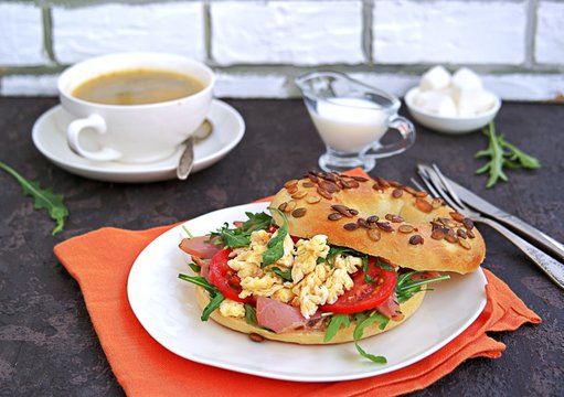 Breakfast, bagel with scramble egg, ham, arugula and vegetables on a white plate on a dark background.