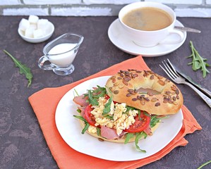 Breakfast, bagel with scramble egg, ham, arugula and vegetables on a white plate on a dark background.