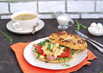 Breakfast, bagel with scramble egg, ham, arugula and vegetables on a white plate on a dark background.