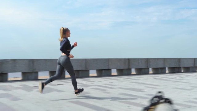 Side view tracking right shot of young female runner jogging on promenade on summer day, full length