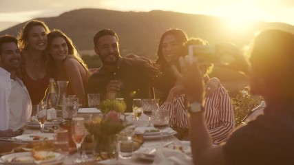 Group of young friends sitting around table and posing while a man taking their picture with her mobile phone. Man taking photographs of friends at dinner party out in the yard. - Powered by Adobe