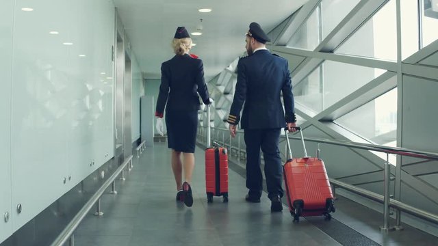 Pilot and stewardess with red suitcases walking by airport. Back view.