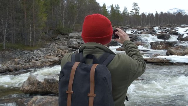 Traveler in a red hat with a backpack full back stopped to take a photo of a mountain river. concept of travel and photography.