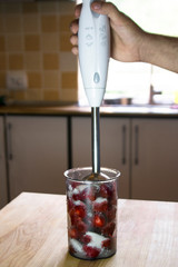 Red ripe strawberries with sugar in a glass. Male hand is holding a blender.