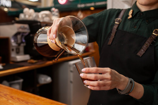 Female barista is pouring black coffee, made in chemex, into a glass, standing in front of coffee ouse interior