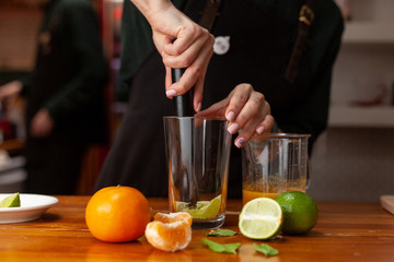 Professional female bartender is making refreshing citrus lemonade kneading lemons with a mortar in a glass, placed on a wooden table