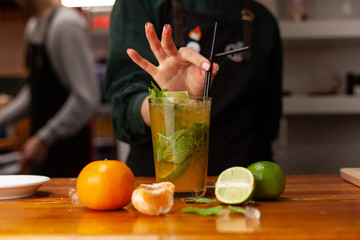 Professional female bartender is making refreshing citrus lemonade behind the bar counter, decorated with ingredients: lime, orange and lemon