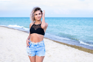 beautiful young woman with curly hairs in denim shorts and swimsuit is happy to be on the beach near the ocean. Ocean and grass on the background