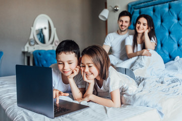 Cute girl and boy lie in bed with laptop watching movie, behind parents in bed at home.
