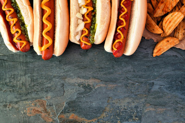 Hot dogs with toppings and potato wedges. Top border, overhead view on a dark stone background with...
