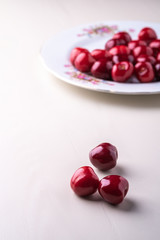 Three cherry berries with white plate with cherries in background on wooden white background angle view