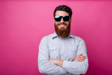 Portrait of smiling man with beard wearing sunglasses and standing over pink background with crossed arms