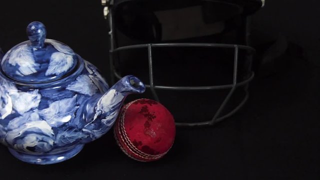 Cricket ball rolls into frame to stop beside a teapot and a cricket helmet.