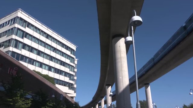 View of modern office buildings from beneath the elevated monorail tracks. Blue train passing overhead following curved tracks away from a tall building. Public transportation.