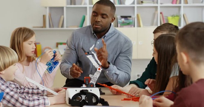 Afican American young male school teacher playing with kids with robots and toys and showing them how to operate them or assemble.