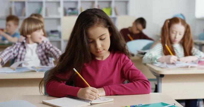 Small beautiful schoolgirl with long dark hair wriying with a pencil an exercise in the copybook during control test at the school lesson.