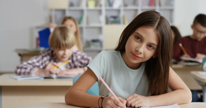 Portrait shot of the Caucasian little teen girl writing in the copybook during test at the lesson in the classroom and smiling to the camera.