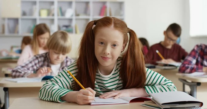 Portrait shot of the Caucasian teen red-haired schoolgirl writing in the copybook at the lesson in the classroom and smiling cheerfully to the camera.