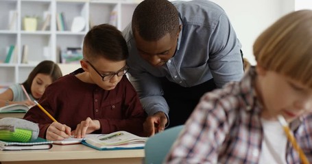 African American young male teacher leaning on the desk in the classroom and explaining something in the textbook to the cute boy.