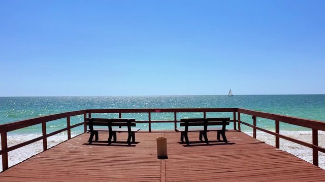 Two wooden benches on a deck facing the beautiful ocean, with waves crashing and a boat moving in the horizon. Clearwater, Florida, USA. (FYI: footage available from a different angle in my portfolio)