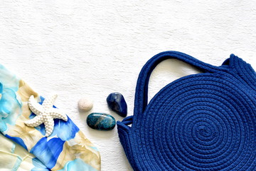 Beach summer fashion concept. Royal blue straw bag, handbag with blue pebbles and turquoise floral silk scarf on the textured white background. Top view, from above. Flat lay, flat-lay. Copy space.