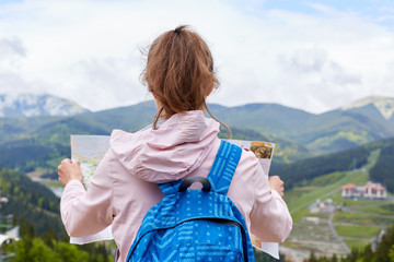 Back view of travel standing at top of hill holding map with current route to move on, looking at magnificent mountain landscape, wearing jacket, blue backpack, having ponytail. Travelling concept.