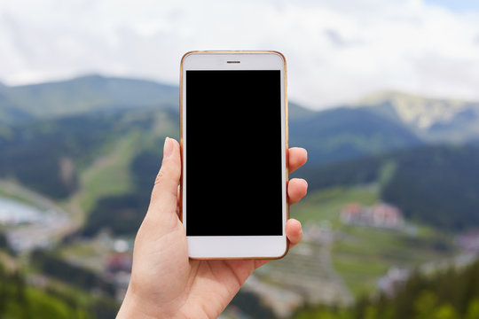Outdoor image of one hand holding and showing white smartphone with blank black desktop screen with blur green mountains on background, mobile phone switched off. Copyspace for advertisement.