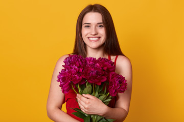 Beauty studio portrait of romantic model woman with burgundy peony flowers. Attractive female posing with toothy smile, embracing her bouquet, expresses happyness and joy. People and presents concept.