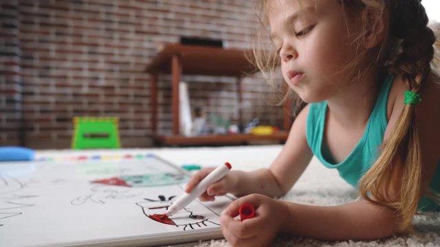 Video Shot of happy little girl lying and drawing with felt-tip pen on white capet at home. Childhood concept. Studio video shot of child's daily life in 4K definition.