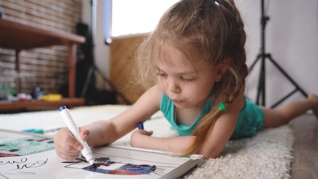 Video Shot of happy little girl lying and drawing with felt-tip pen on white capet at home. Childhood concept. Studio video shot of child's daily life in 4K definition.