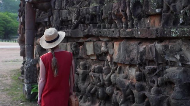 Caucasian woman in red dress is exploring amazing bas-reliefs of the Leper King terrace located in the northwest corner of the Royal Square of Angkor Thom, Cambodia