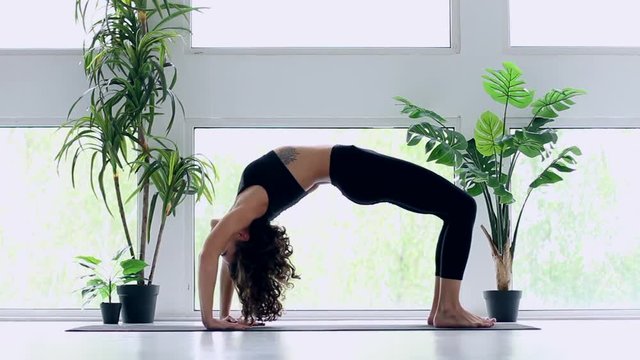 Young slender female is doing eka pada urdhva dhanurasana. Athletic brunette woman in black uniform is practicing yoga, balancing in one legged wheel pose at fitness studio. Healthy lifestyle concept.