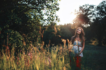 Fototapeta na wymiar Young pregnant woman in the park outdoors. Calm pregnant woman in third trimester. Walking in public garden. Sunset in the forest