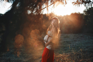 Fototapeta premium Young pregnant woman in the park outdoors. Calm pregnant woman in third trimester. Walking in public garden. Sunset in the forest
