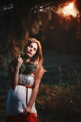 Fototapeta premium Young pregnant woman in the park outdoors. Calm pregnant woman in third trimester. Walking in public garden. Sunset in the forest