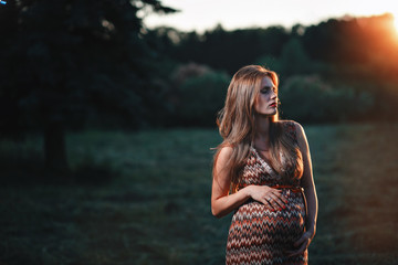 Young pregnant woman in the park outdoors. Calm pregnant woman in third trimester. Walking in public garden. Sunset in the forest