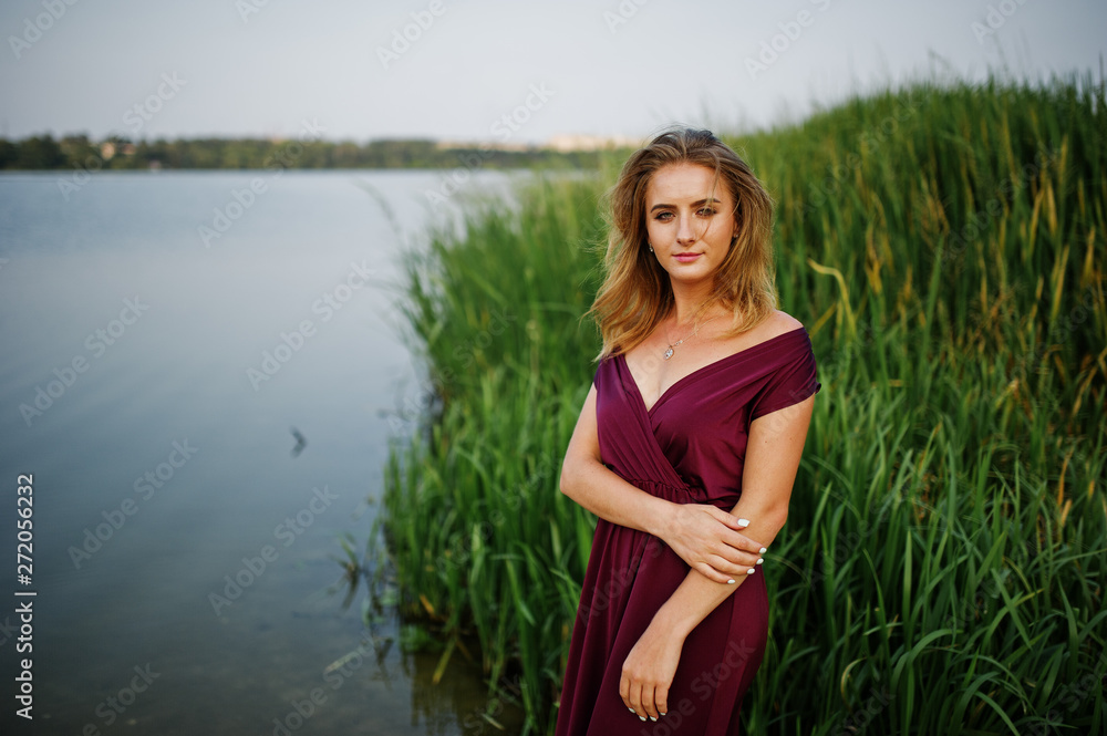 Wall mural Blonde sensual woman in red marsala dress posing against lake with reeds.