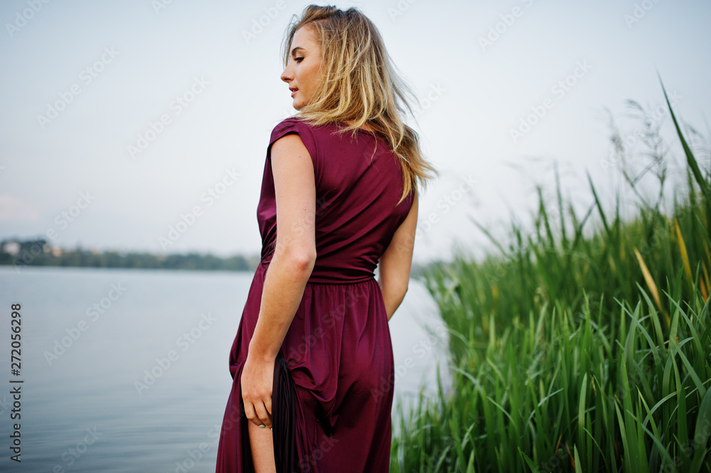 Canvas Prints Blonde sensual woman in red marsala dress standing in water of lake with reeds and show her nude leg.