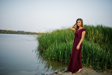 Obraz premium Blonde sensual barefoot woman in red marsala dress posing against lake with reeds.