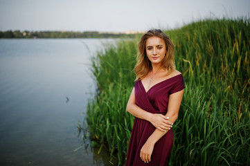 Fototapeta premium Blonde sensual woman in red marsala dress posing against lake with reeds.
